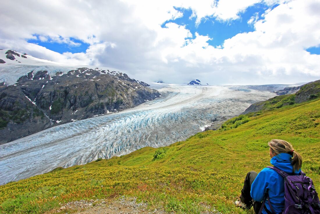 Blonde female traveller sits on a hill overlooking Exit Glacier in Harding Ice Field