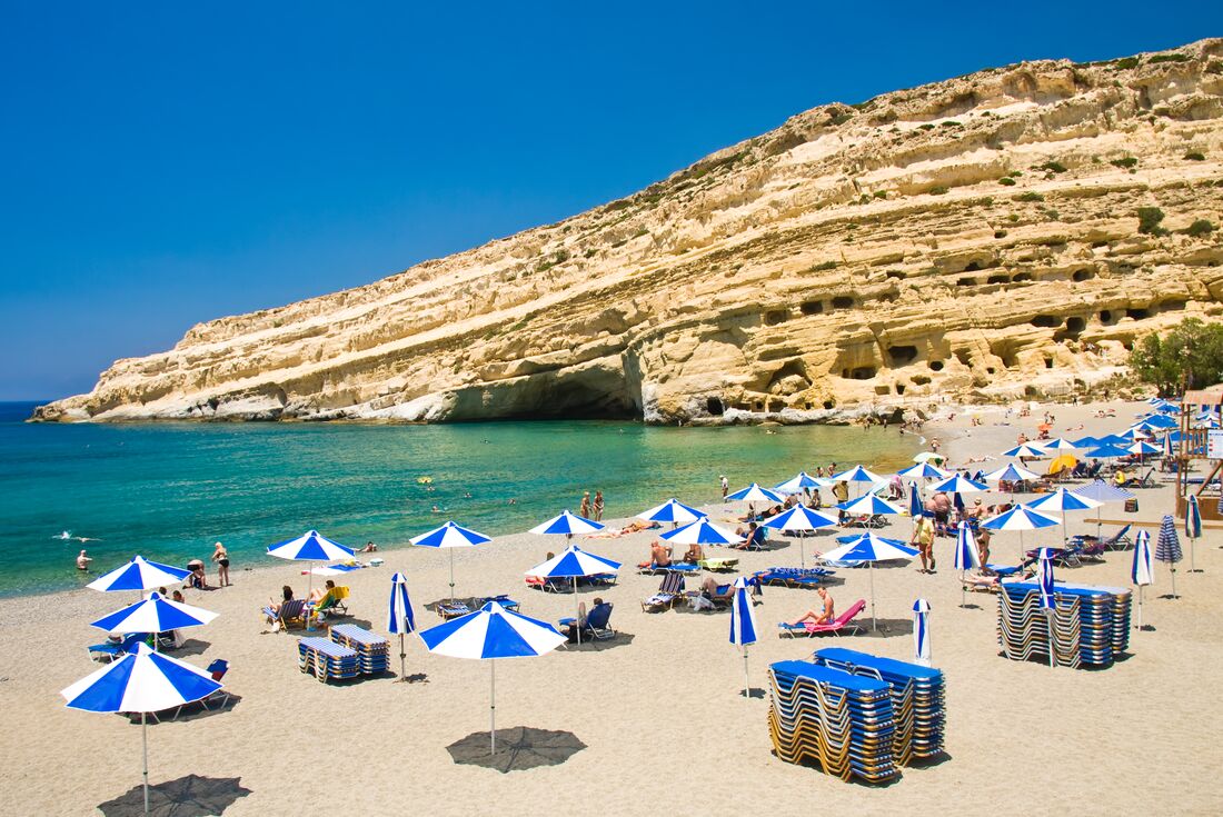 Travellers on the beach with blue and white parasols Matala beach with cliff in background, Crete, Greece
